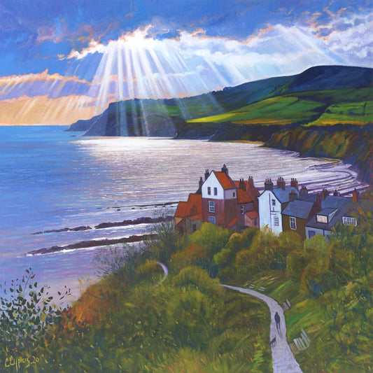 Features the famous headland view over the stunning Robin Hoods Bay, Northeast Coast UK.  The sun breaks through clouds along the coastline the sun's rays reflecting on the surface of the water. A figure walks down the path towards the town