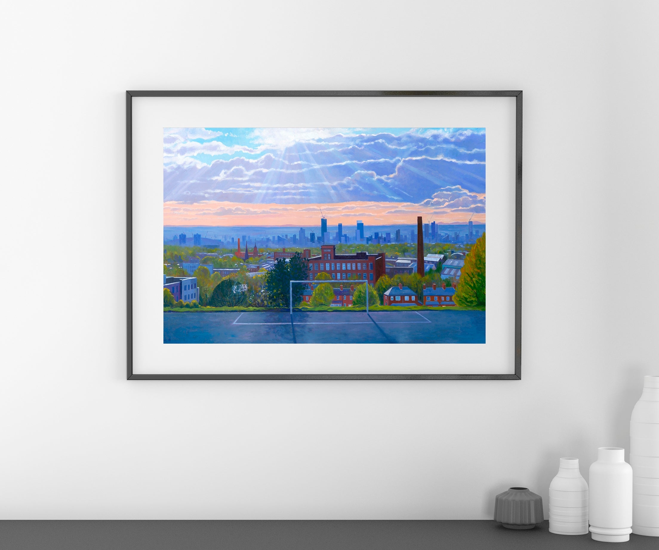 A framed art print depicting a colourful city skyline of Manchester UK at sunset, hung on a white wall above a dark gray surface with decorative white vases.