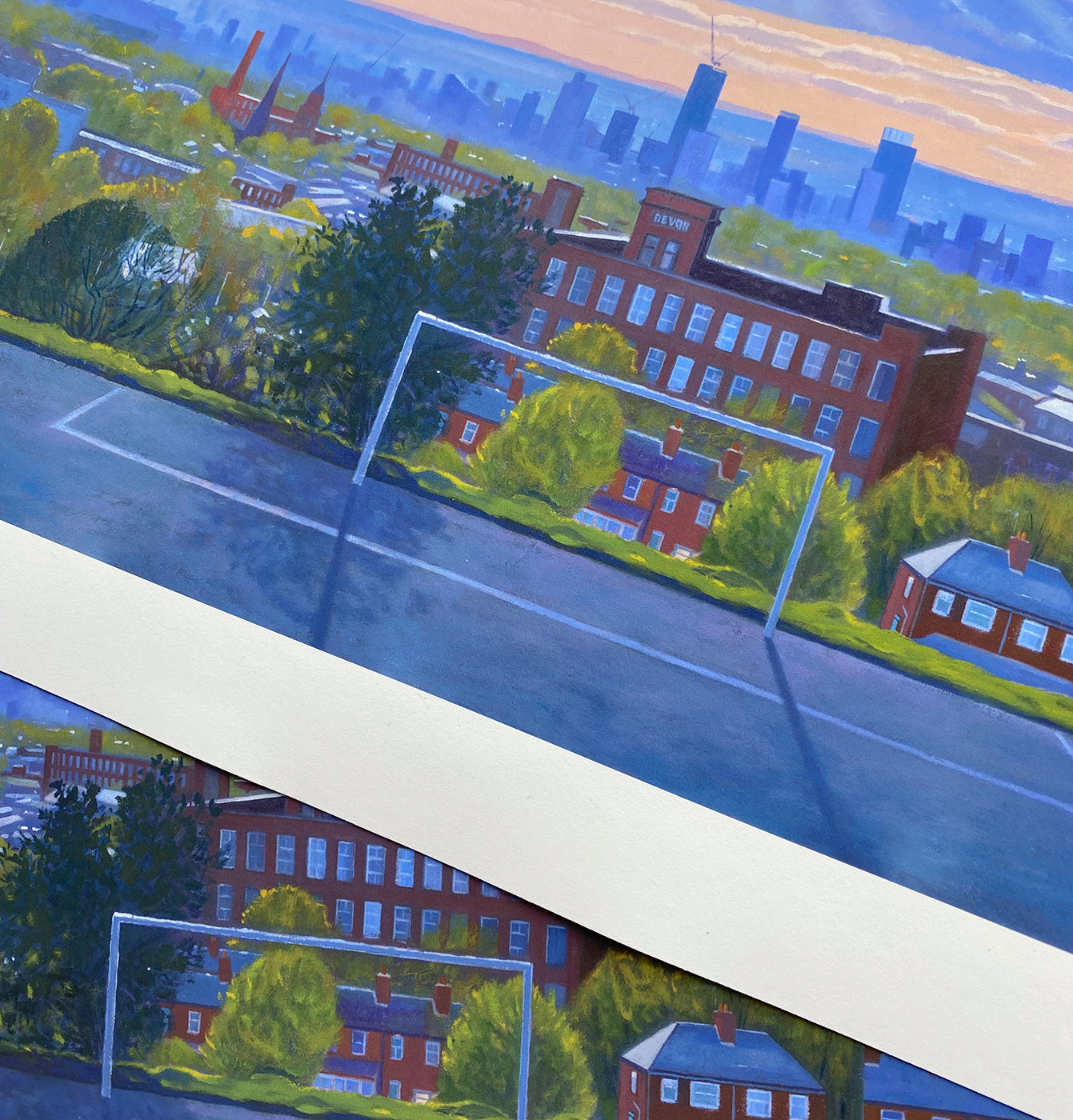A square art print or painting depicting a suburban street scene with brick houses, green trees, a blue road, and a distant city skyline of Manchester under a warm sky. The print is shown at an angle with a white border visible.