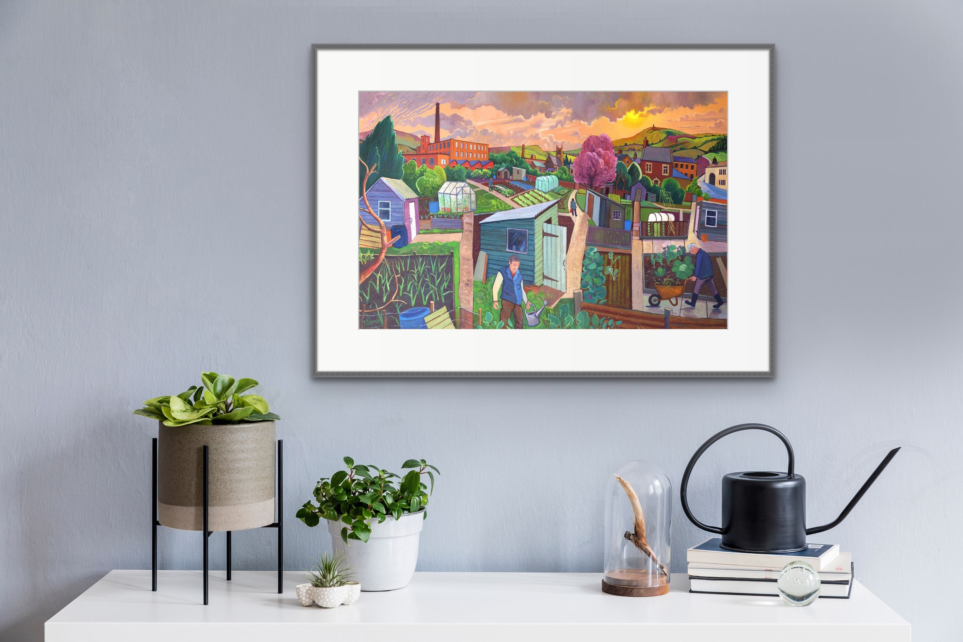 Allotted Time Print displayed in a frame hung on the wall. A detailed scene combining elements of beautiful landscape surrounding an allotment,  plot holders tending to their prize vegetables and flowers on a summers day at sunrise.  
