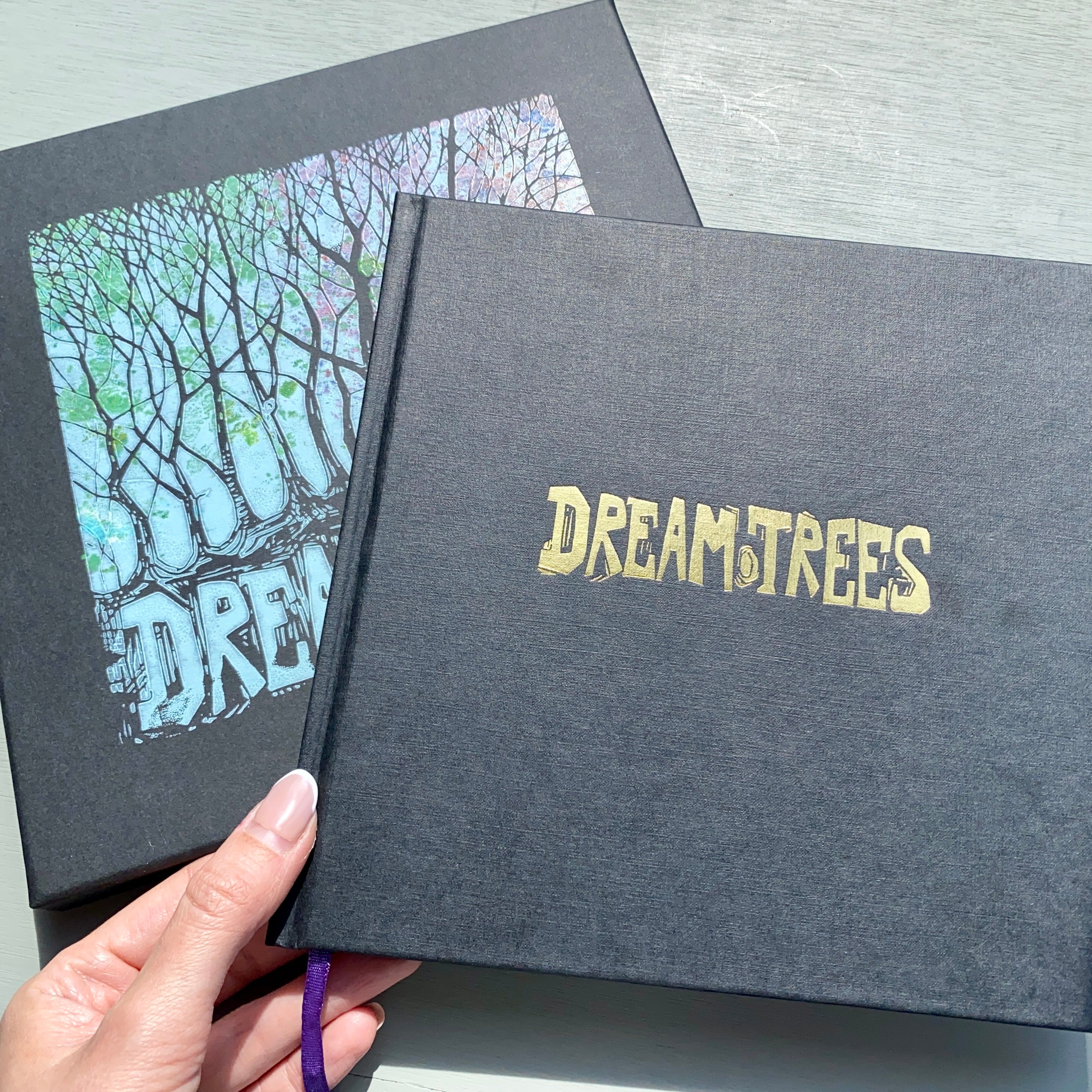 Limited edition hardback book titled 'DREAM TREES' by artist Chris Cyprus with a black cover featuring gold embossed text. A hand is holding the book open to show the title page with a linocut print.