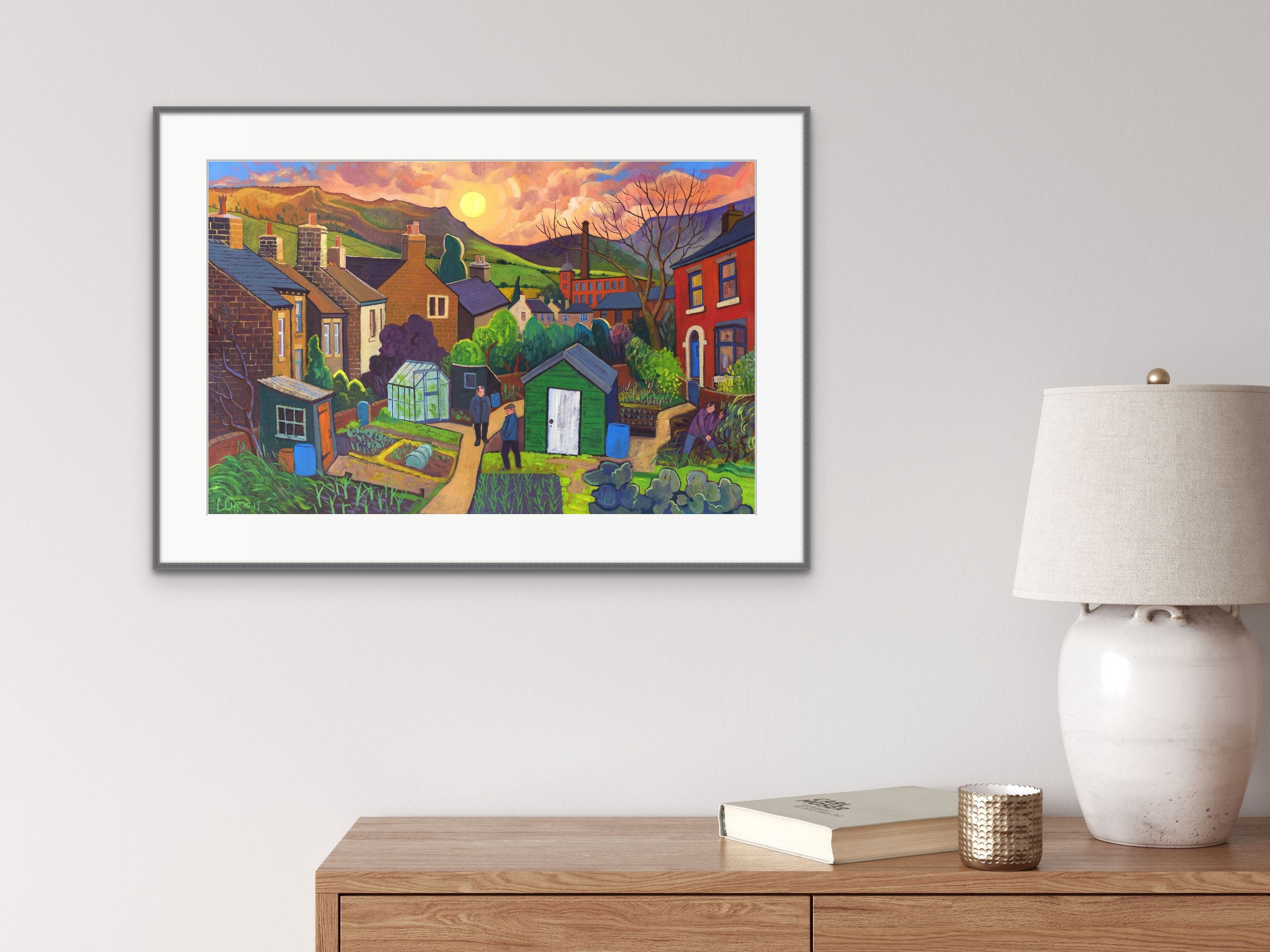 Garden Conversations Print shown displayed  in a frame on the wall with a cabinet and lamp at the side. 
